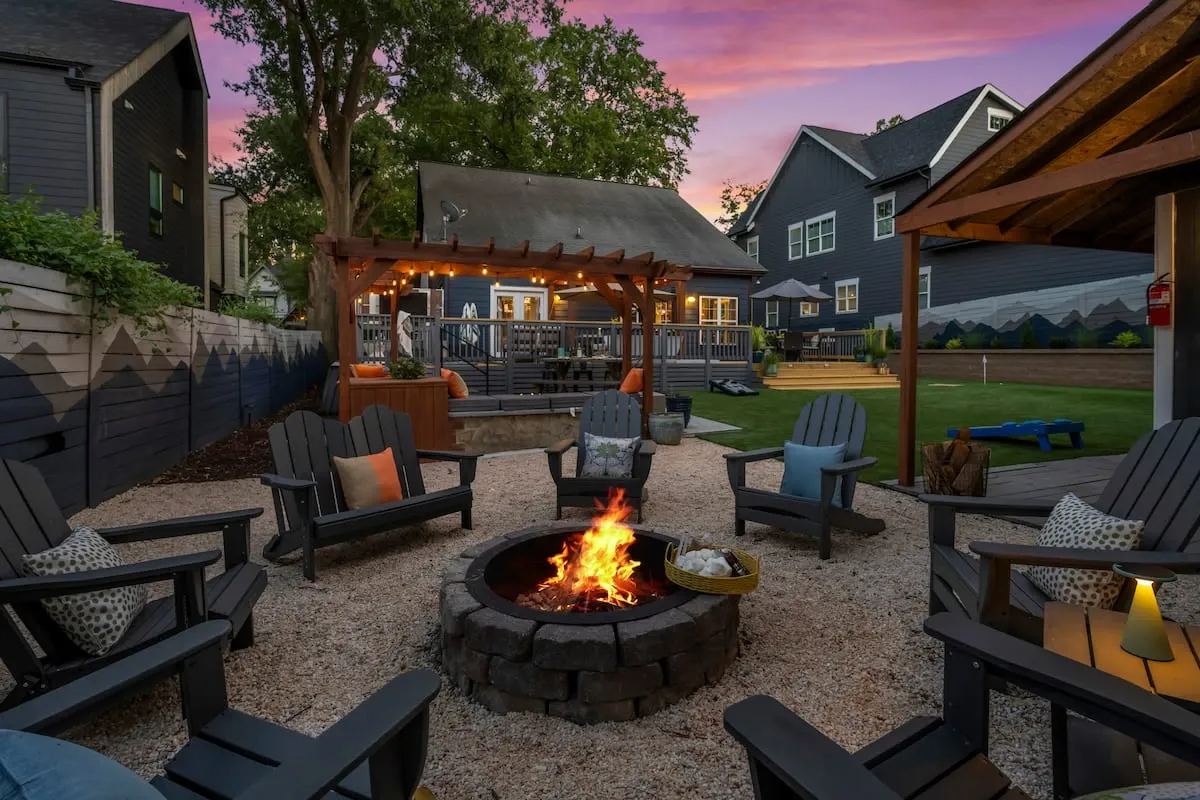 An inviting outdoor living area and backyard fire pit at a rental property, featuring Adirondack chairs, lawn games, and a deck with string lights at sunset.