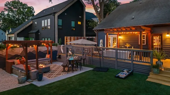 A modern backyard at dusk featuring a wooden deck with string lights and an umbrella, a lower patio area with a fire pit and seating, and a green lawn with a cornhole board.